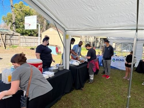People serving food under a tent at an outdoor event with tables and a banner.