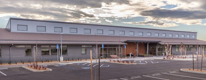 A modern, single-story building with a flat roof, large windows, and an empty parking lot under a partly cloudy sky.