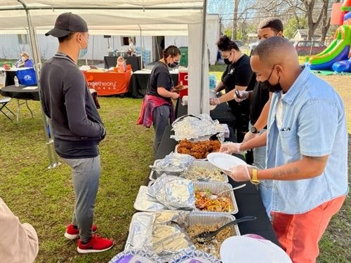 People serving and receiving food at an outdoor event under a canopy, with tables and a bouncy castle in the background.