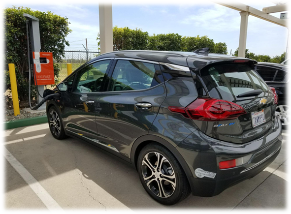 A gray Chevrolet Bolt EV is parked at a charging station with a cable plugged in.