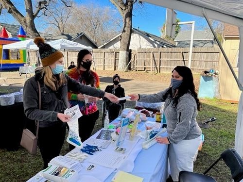 People at an outdoor booth exchanging papers. There are informational materials and products on the table.