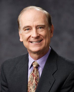 A man in a suit and tie, smiling, with a dark gray background.
