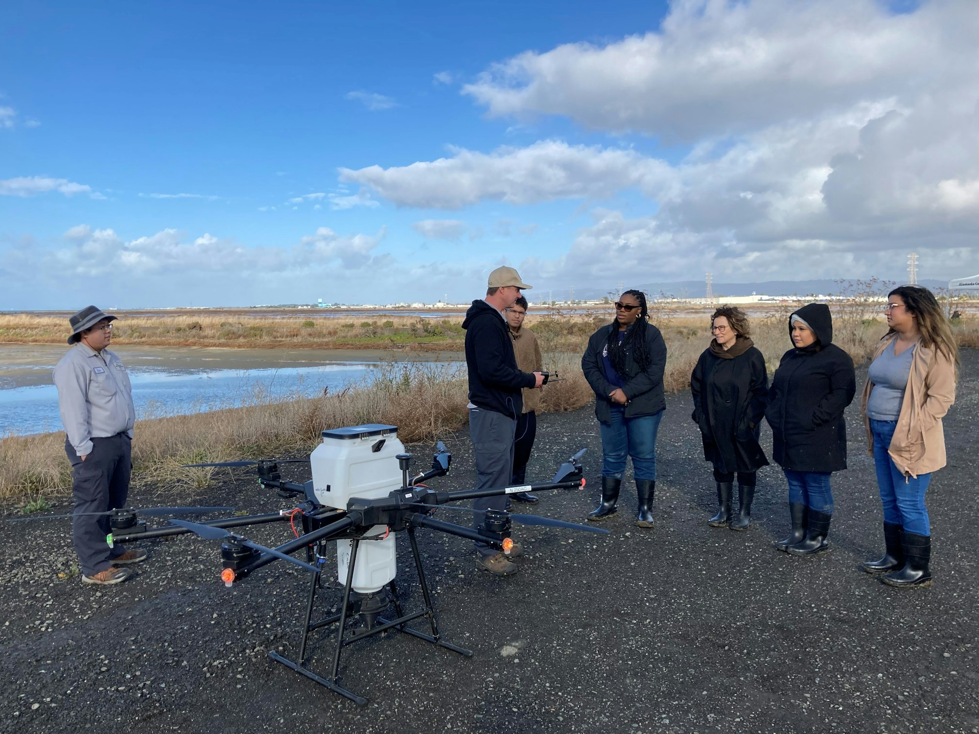 A group of people standing outdoors, observing a large drone on a paved area near a body of water.