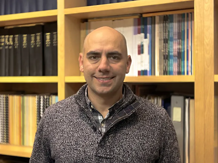 A smiling man stands in front of a wooden bookshelf filled with various books and notebooks.