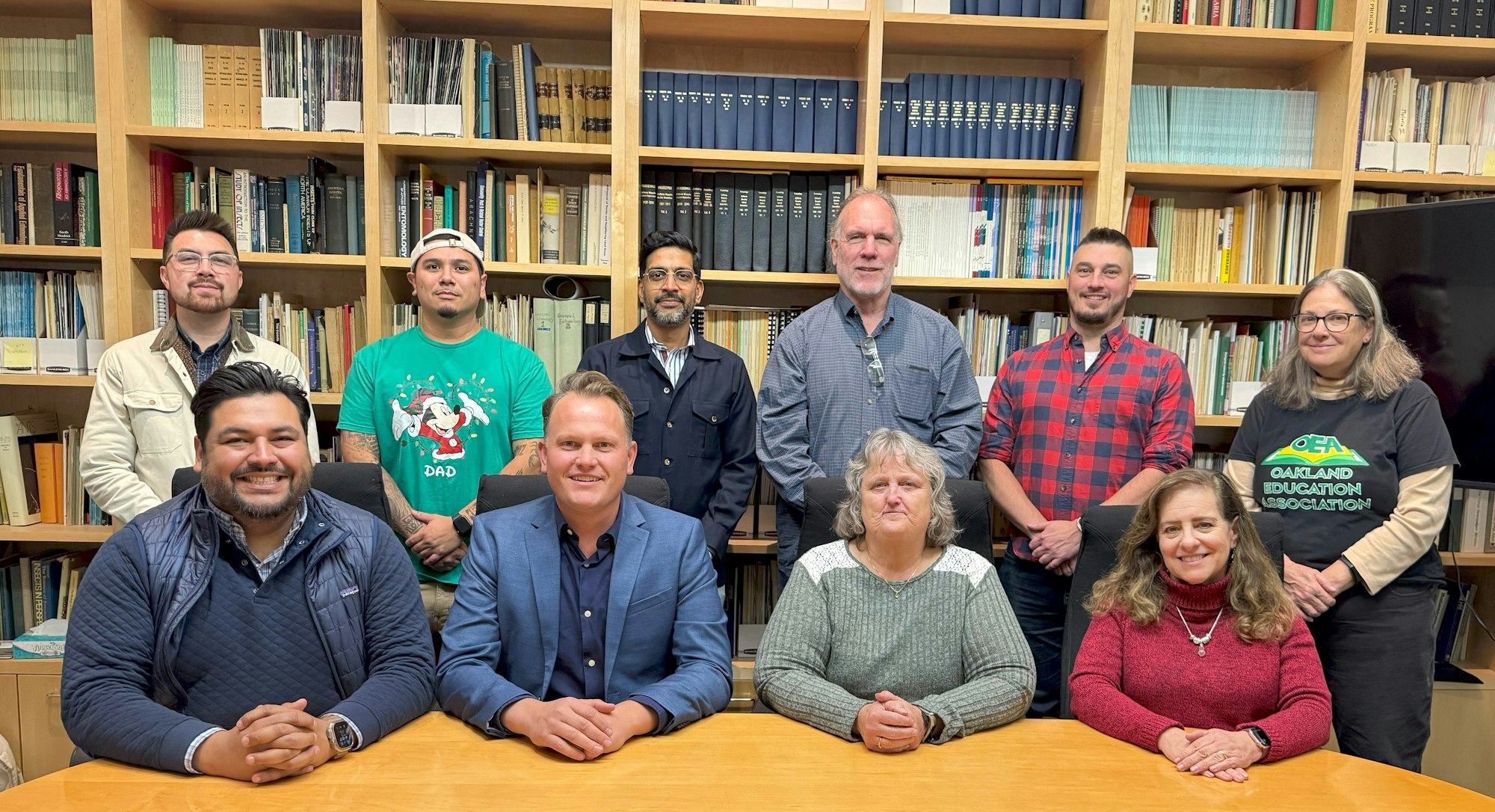 A group of people in a library setting, some seated, some standing, with bookshelves in the background.