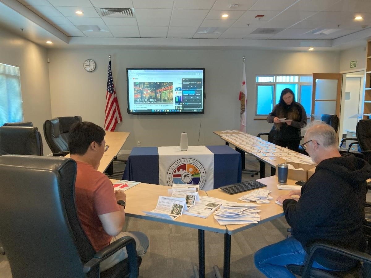 People in a meeting room with a screen displaying content, flags, flyers on a table, and a seal on the podium.