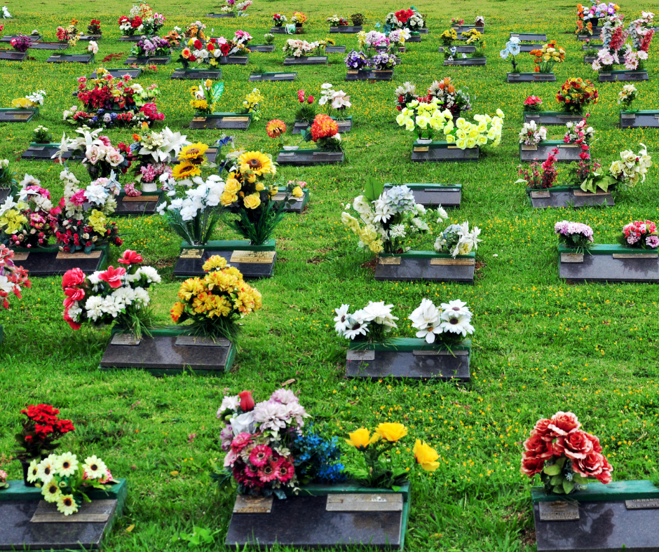 The image shows a cemetery with numerous graves adorned with colorful artificial flowers, set on green grass.