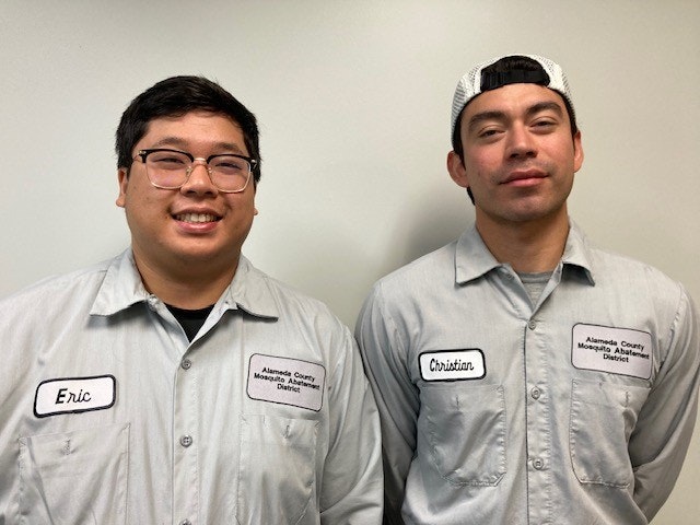 Two people smiling, wearing gray uniforms with name tags, in front of a plain background.