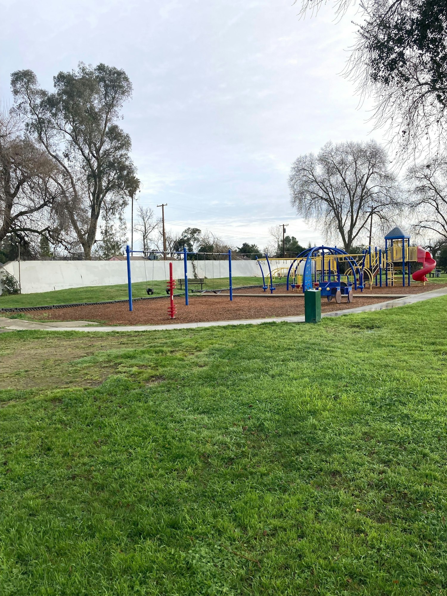 A playground with colorful equipment, a slide, swings, and a climbing structure, surrounded by grass and trees.