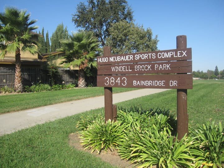 Sign for Hugo Neubauer Sports Complex and Windell Brock Park at 3843 Bainbridge Dr., surrounded by greenery and palm trees.