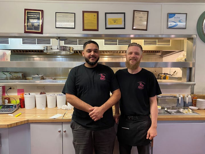 Two smiling men in black t-shirts standing in a restaurant kitchen with awards on the wall behind them.