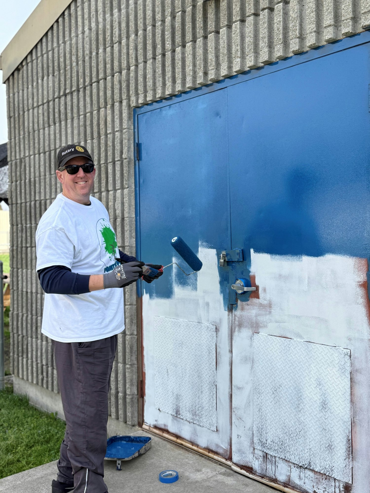 A person in a Rotary hat paints a metal door blue with a roller, smiling, next to paint supplies.