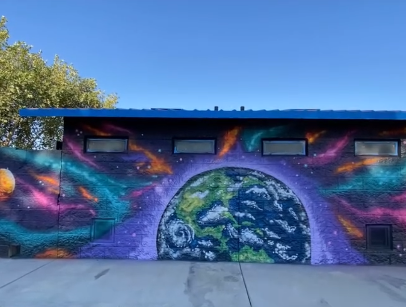 A colorful mural on a building wall depicting a vibrant cosmic scene with Earth, planets, and swirling galaxies.
