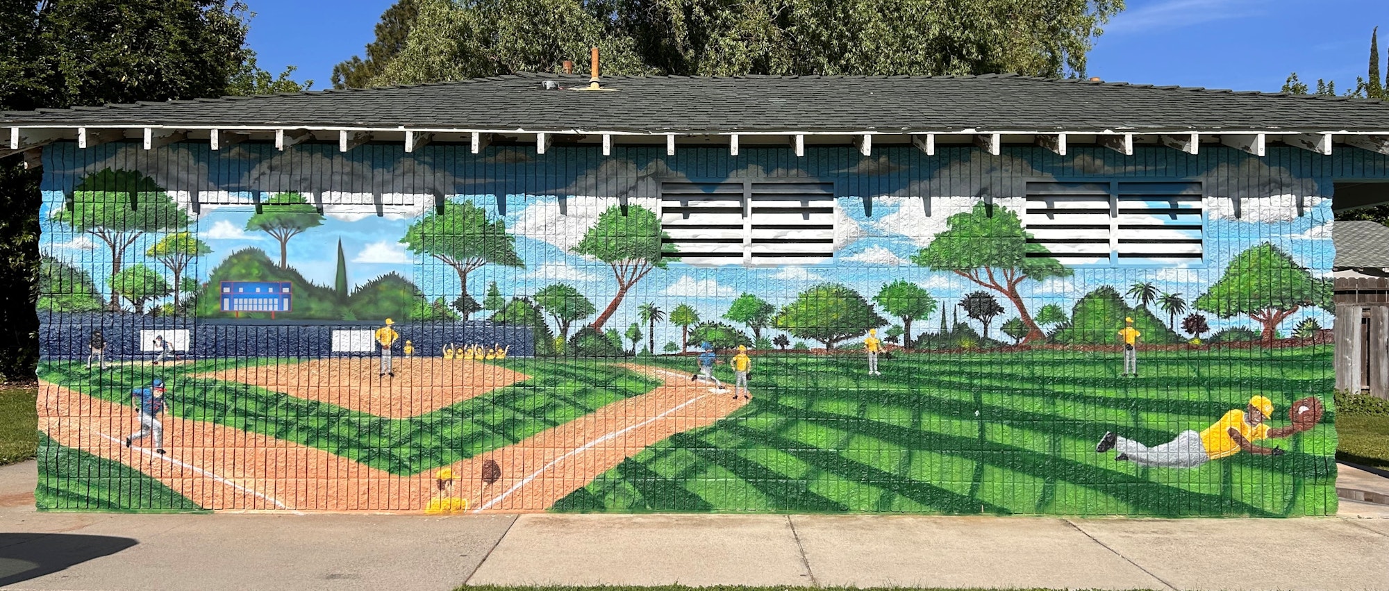 A vibrant mural of baseball players in a lush park, with trees and clouds in the background, on a building wall.