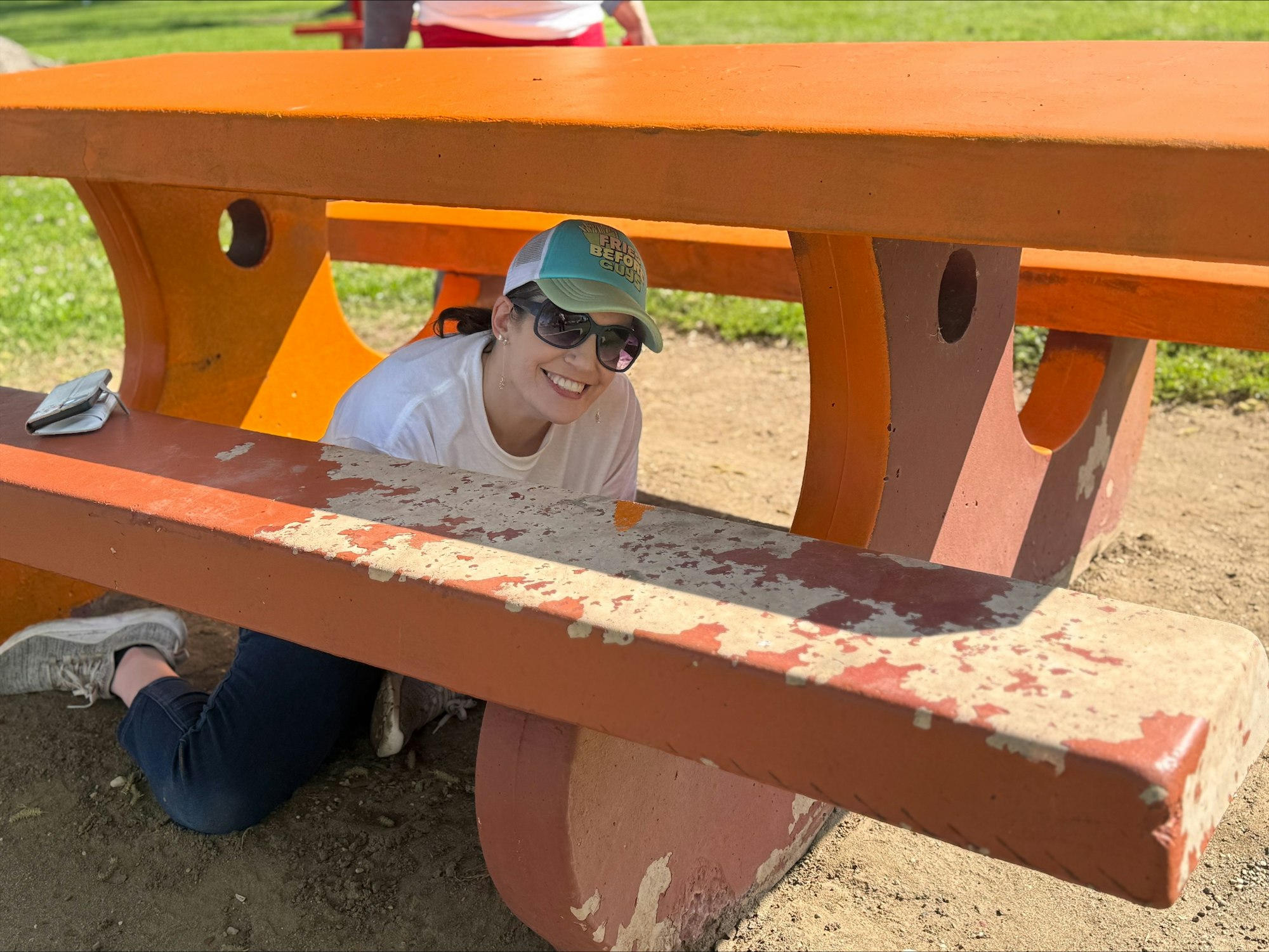 A person smiling, crouched under an orange picnic table in a park, wearing sunglasses and a cap.
