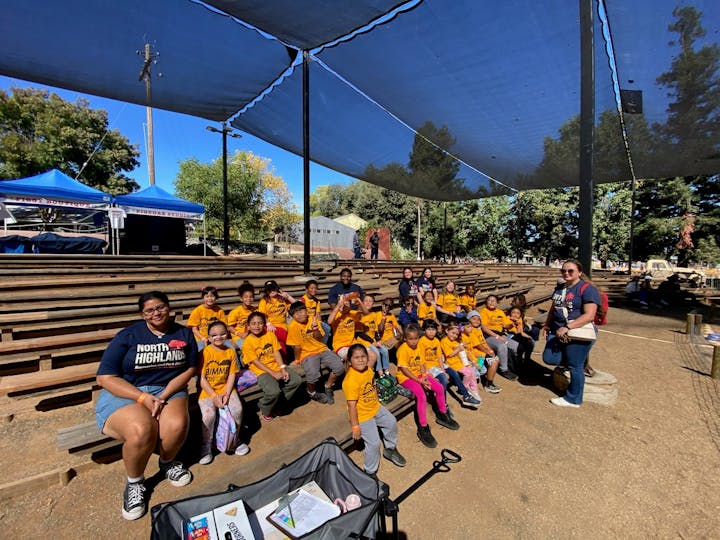 A group of children in matching yellow shirts poses for a photo at an outdoor venue, with benches and tents in the background.