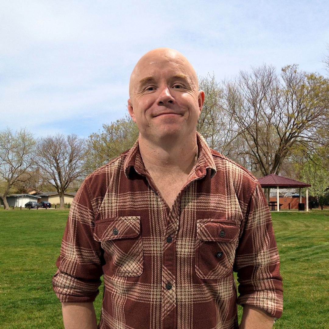 A person in a plaid shirt stands smiling in a park with trees, grass, and a gazebo in the background.