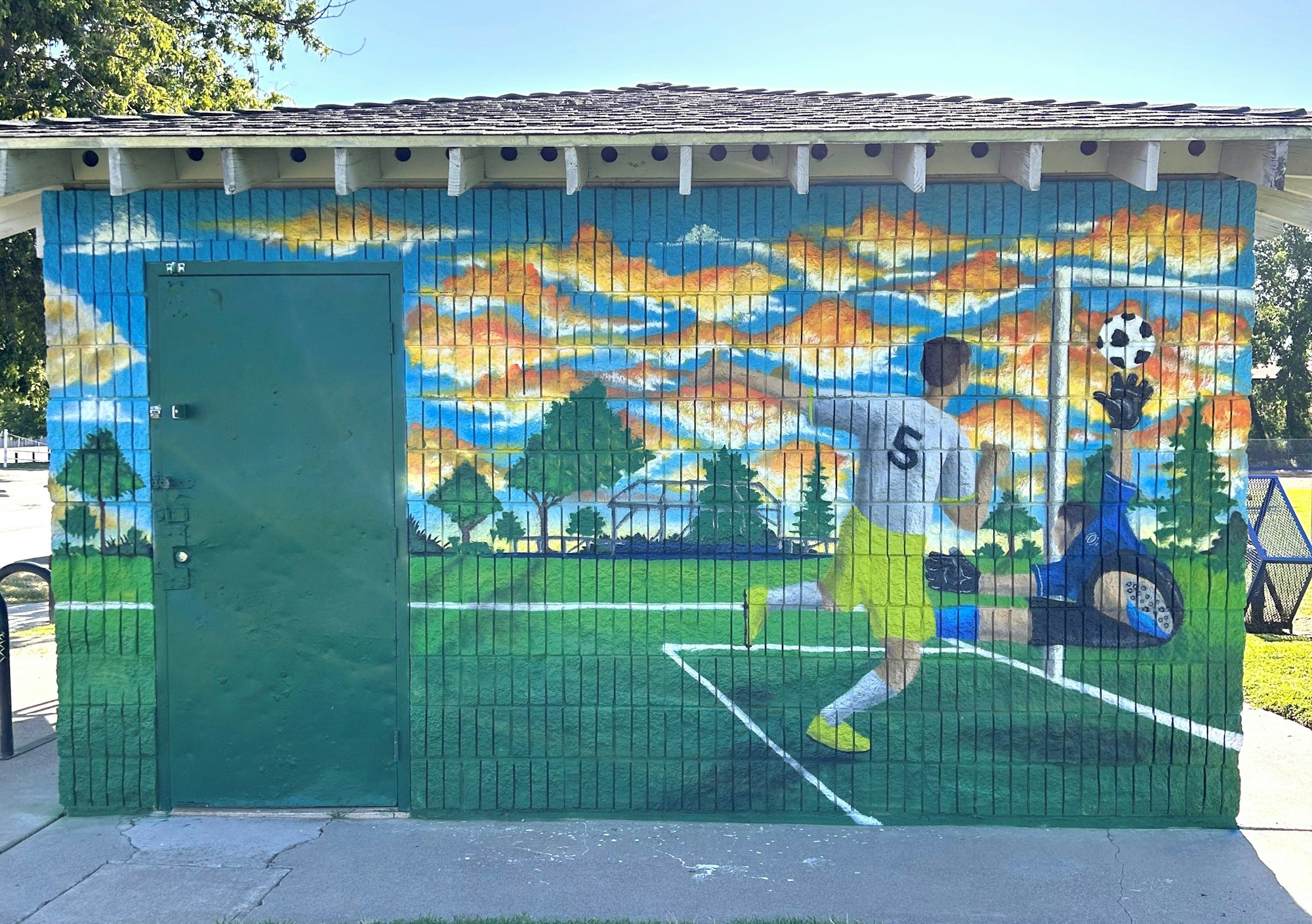 Mural of a soccer player scoring a goal against a goalie, with a scenic background of trees and a sunset sky.