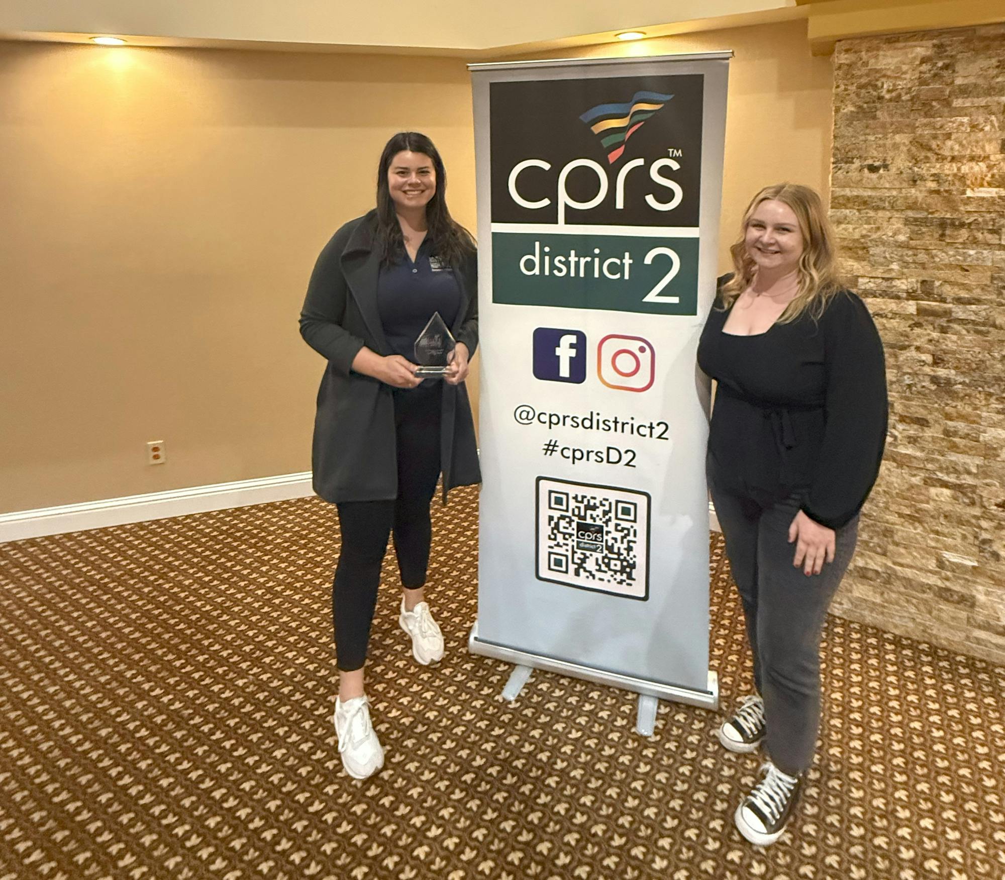 Two people standing beside a CPRS District 2 banner; one is holding an award.