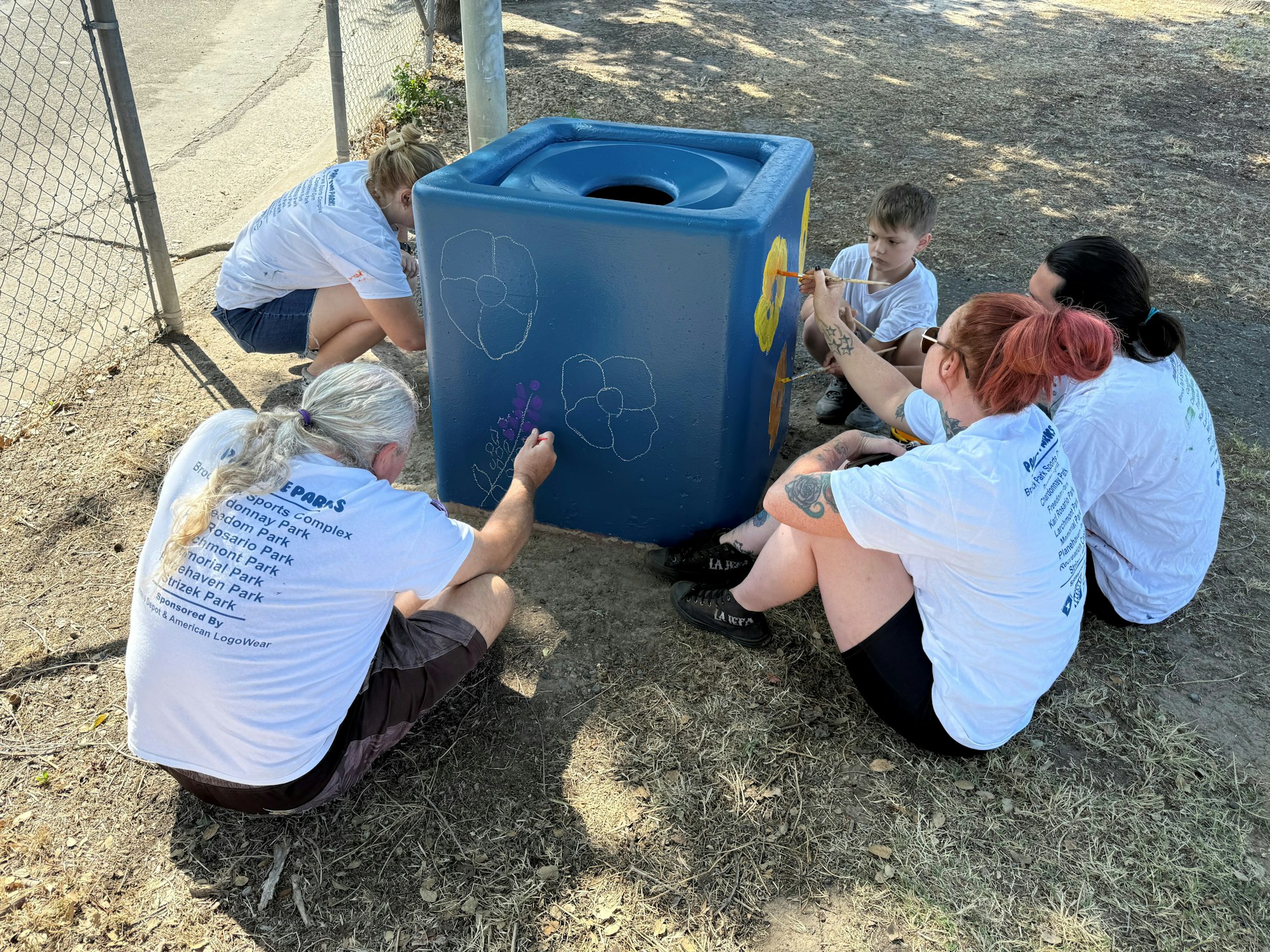 People painting a blue trash bin with flowers, sitting on the ground wearing white shirts.