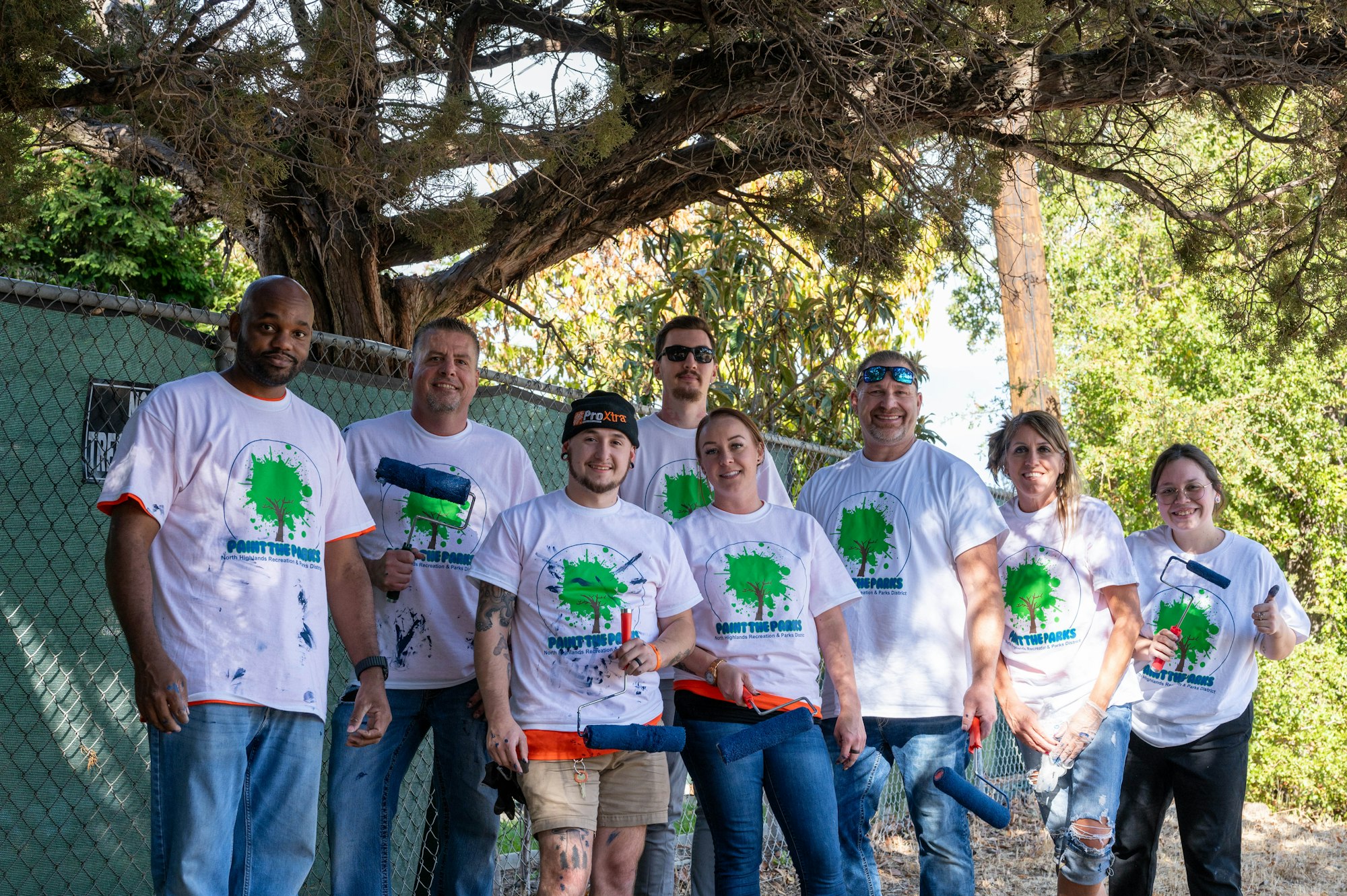 A group of people in matching T-shirts holding painting supplies outdoors.