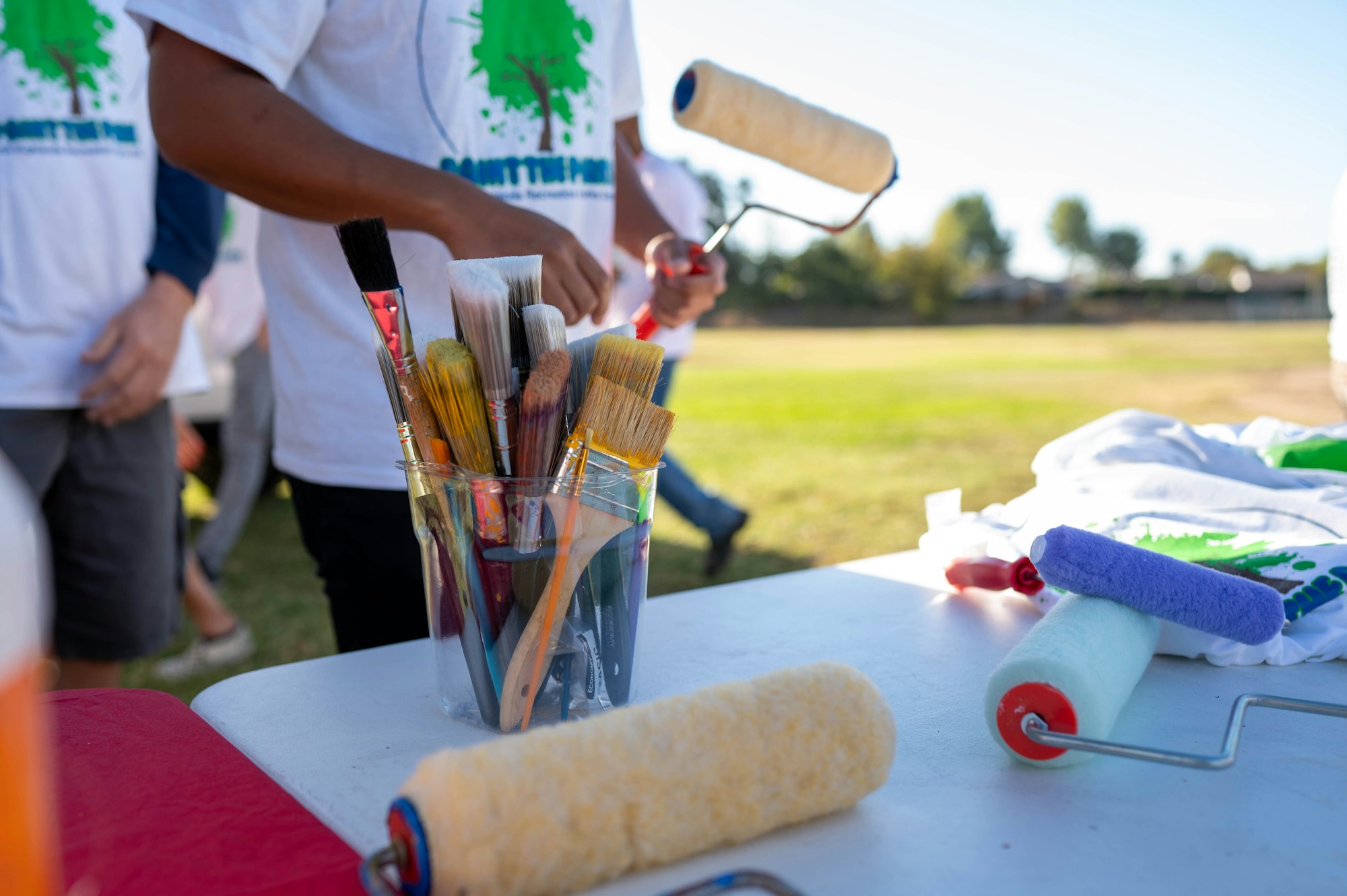 Paintbrushes in a glass jar and paint rollers on a table with people in the background, possibly at a community event.