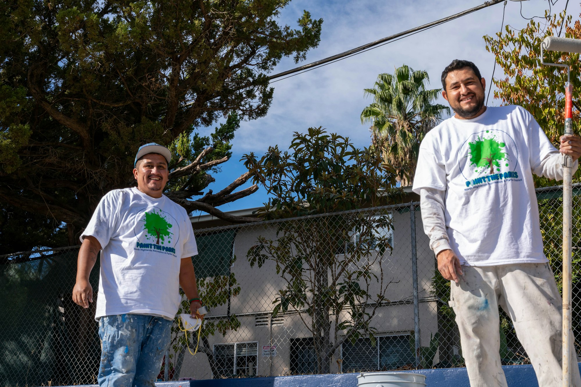 Two smiling individuals in paint-splattered clothing with rollers, standing by a fence, trees and a blue sky in the background.