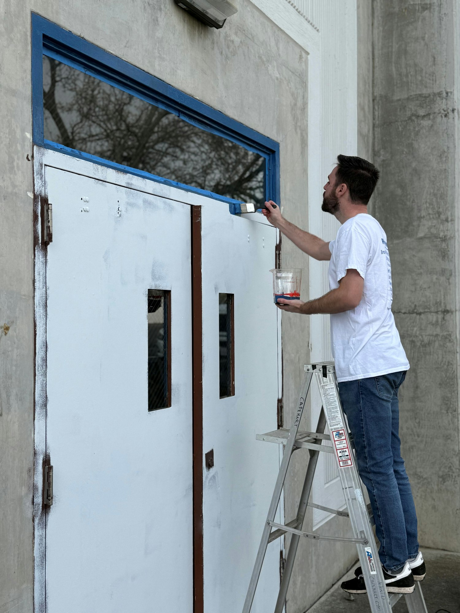 Person on a ladder painting a door frame blue with a brush.