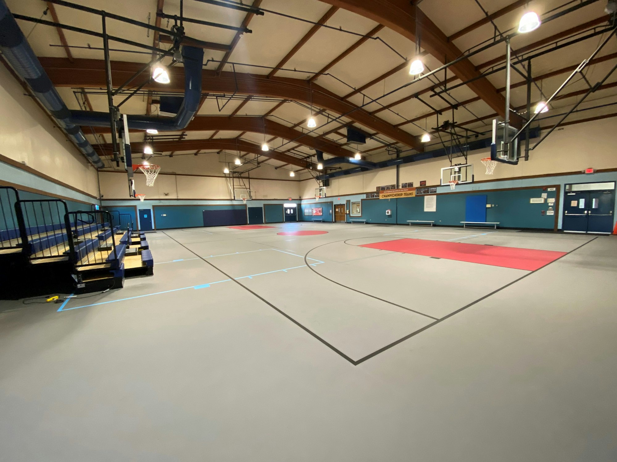 An indoor basketball court featuring hoops, seating, and a gray playing surface with red and blue markings.