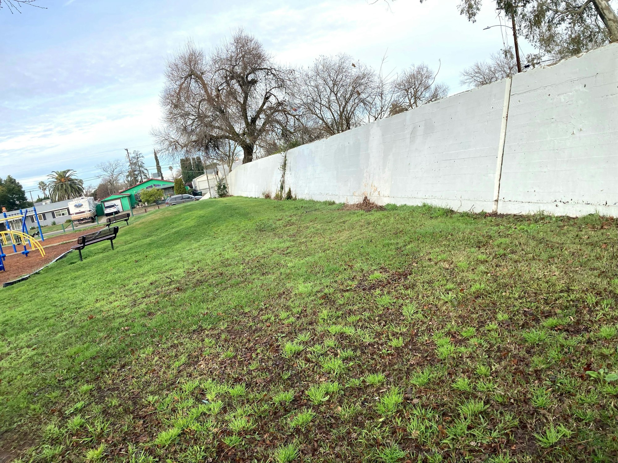 A grassy park with a bench, playground equipment, trees, and a tall white wall under a cloudy sky.