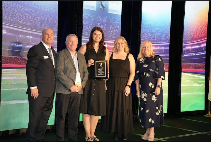 Five people standing on a stage; one holds a plaque. They are indoors with a sports stadium backdrop.