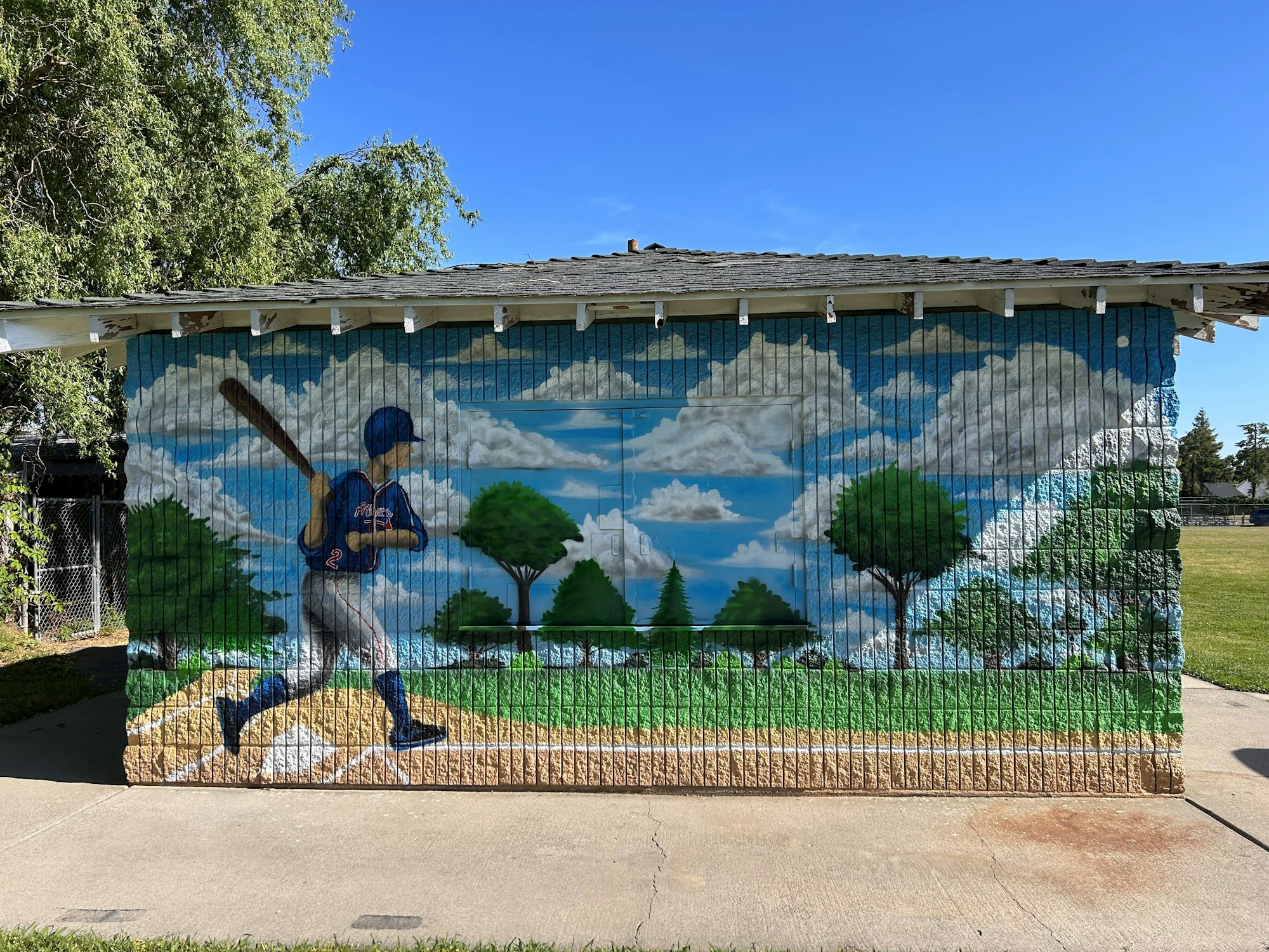 A mural of a baseball player swinging a bat, set against a backdrop of trees and clouds on a small building.