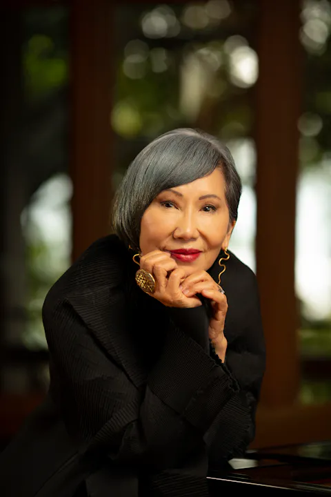 An elegant person with grey hair, wearing black clothes and gold jewelry, posing with a chin rest.