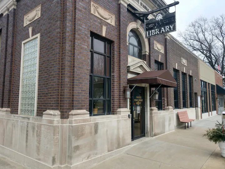 The image shows an exterior view of a public library building with a sign, large windows, and a welcoming entrance.
