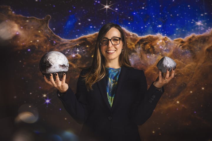 A person holds a globe of Pluto and a small asteroid model, set against a colorful cosmic background.
