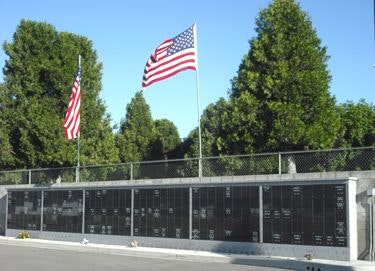 The image shows a memorial wall with engraved names and two American flags, set against a backdrop of trees.