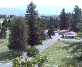 The image shows a cemetery with green lawns, trees, and some buildings in the background.