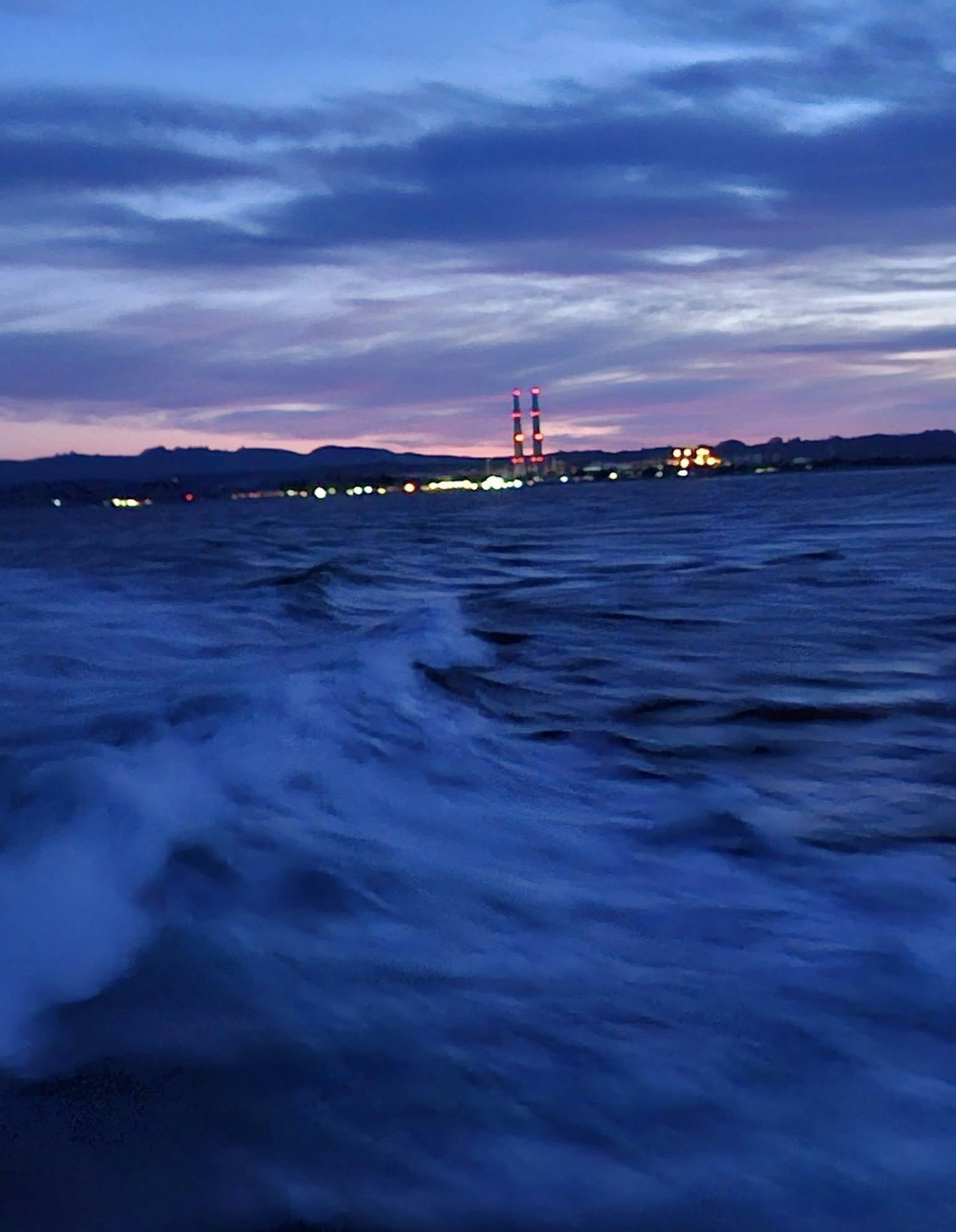 A calm sea at dusk with rolling waves, illuminated by distant lights from structures and a colorful sky.