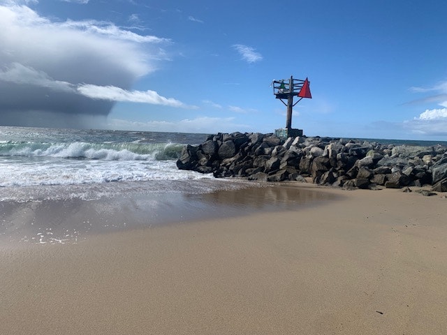 A beach scene with waves, rocky jetty, and a warning flag on a pole against a clear sky with some clouds.
