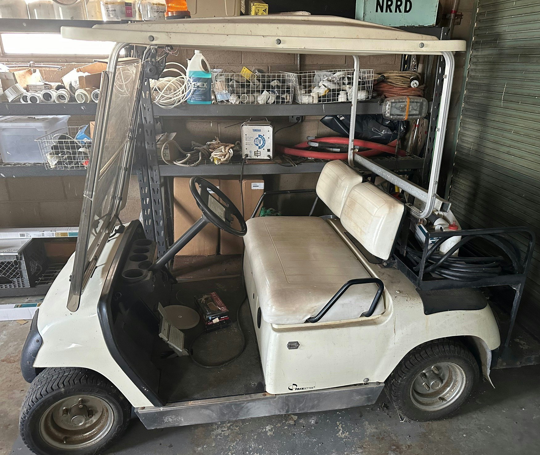 A white golf cart is parked in a cluttered garage with shelves holding various tools and supplies.