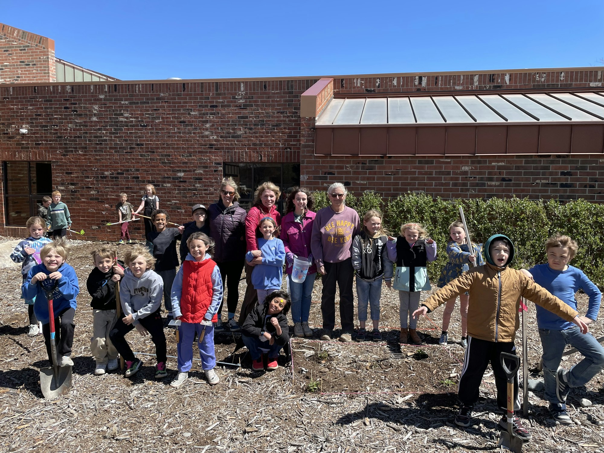 A group of children and adults with gardening tools posing outside in front of a brick building.