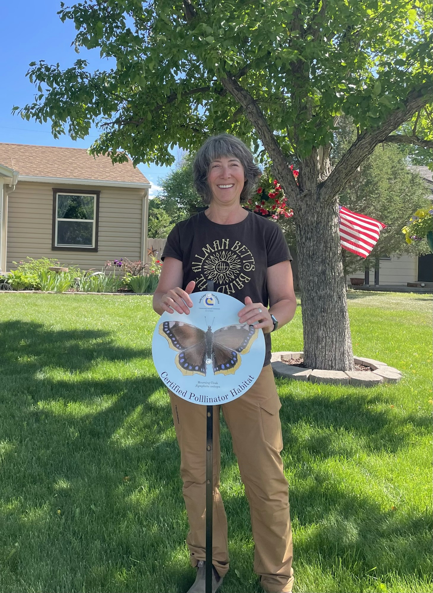 Person holding a "Certified Pollinator Habitat" sign, standing in a sunny garden with an American flag and a tree in the background.