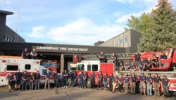 Group of firefighters in uniform in front of fire trucks and station.