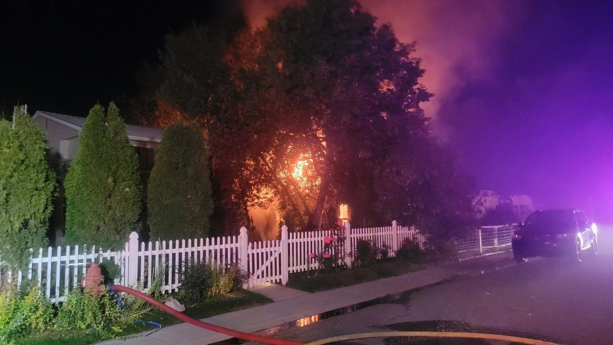 House fire at night with trees, white picket fence, and a fire hose across the street.