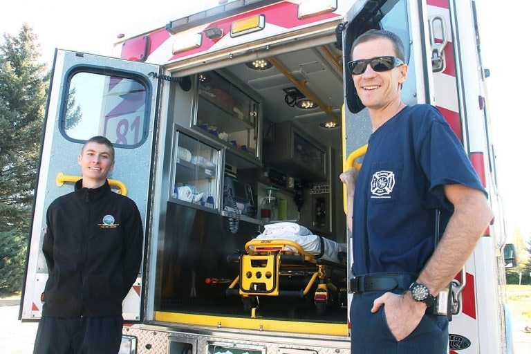 Two paramedics by an ambulance, smiling, emergency medical equipment visible inside.