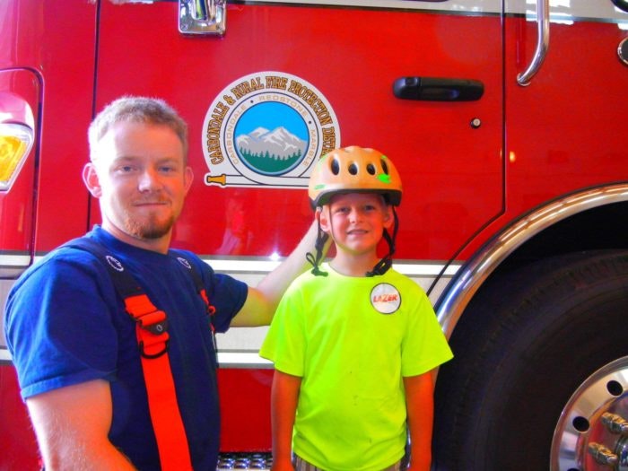 Two individuals are posing in front of a red firetruck; one is a child wearing a helmet and the other is an adult.