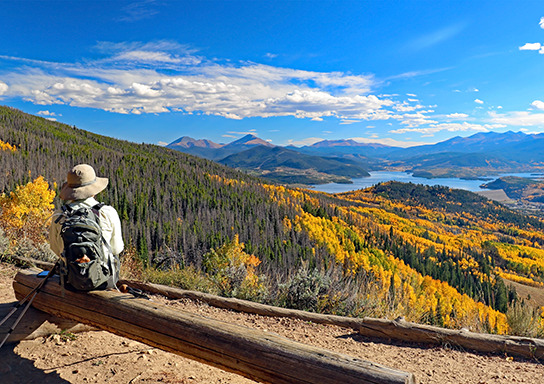 A hiker enjoys a scenic overlook of autumn-colored trees, a lake, and distant mountains under a blue sky.