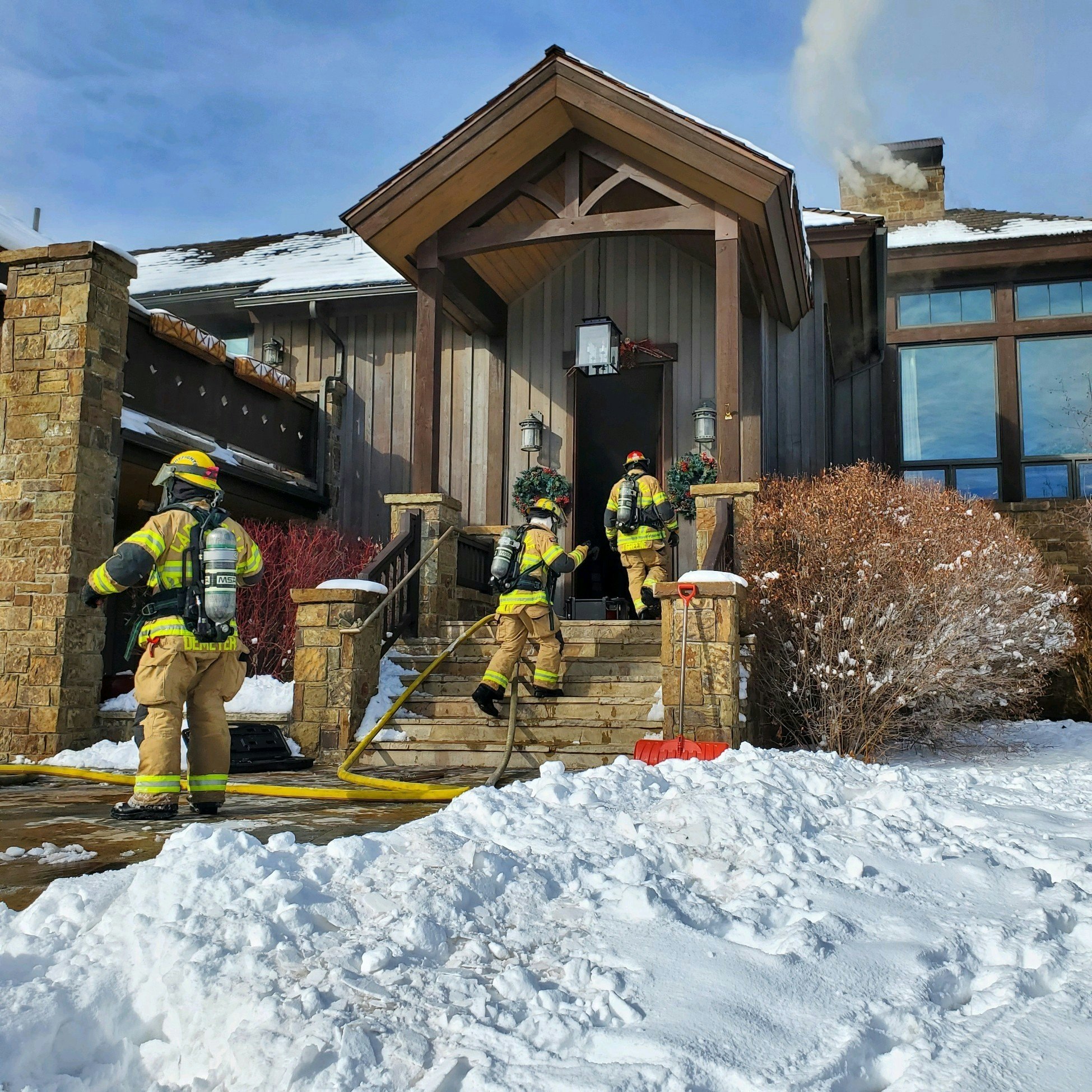 Firefighters in gear outside a snowy house with smoke coming from the chimney.