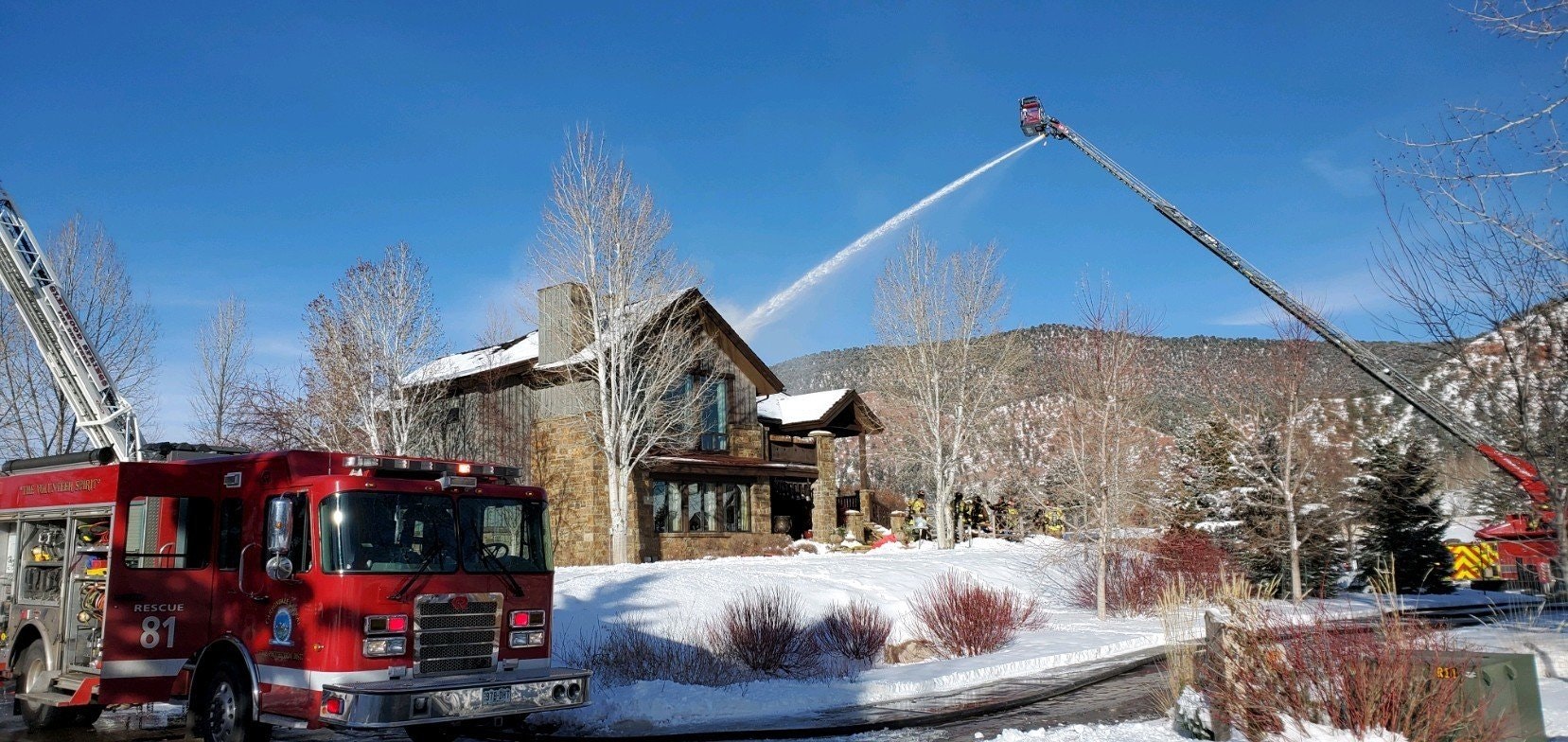 Firefighters using a ladder truck to douse a house with water in a snowy landscape.