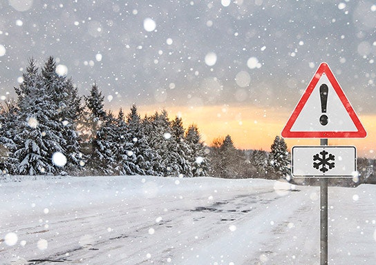 A snowy scene with a warning road sign indicating icy conditions ahead, surrounded by trees under a cloudy sunset sky.
